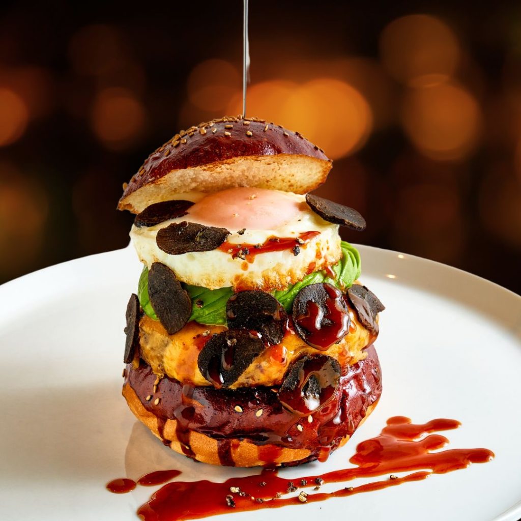 A Halal gourmet burger on a white plate with a dark background. The burger has a black bun, a beef patty, melted cheese, avocado, a fried egg, and black truffle shavings. It is drizzled with a dark sauce.