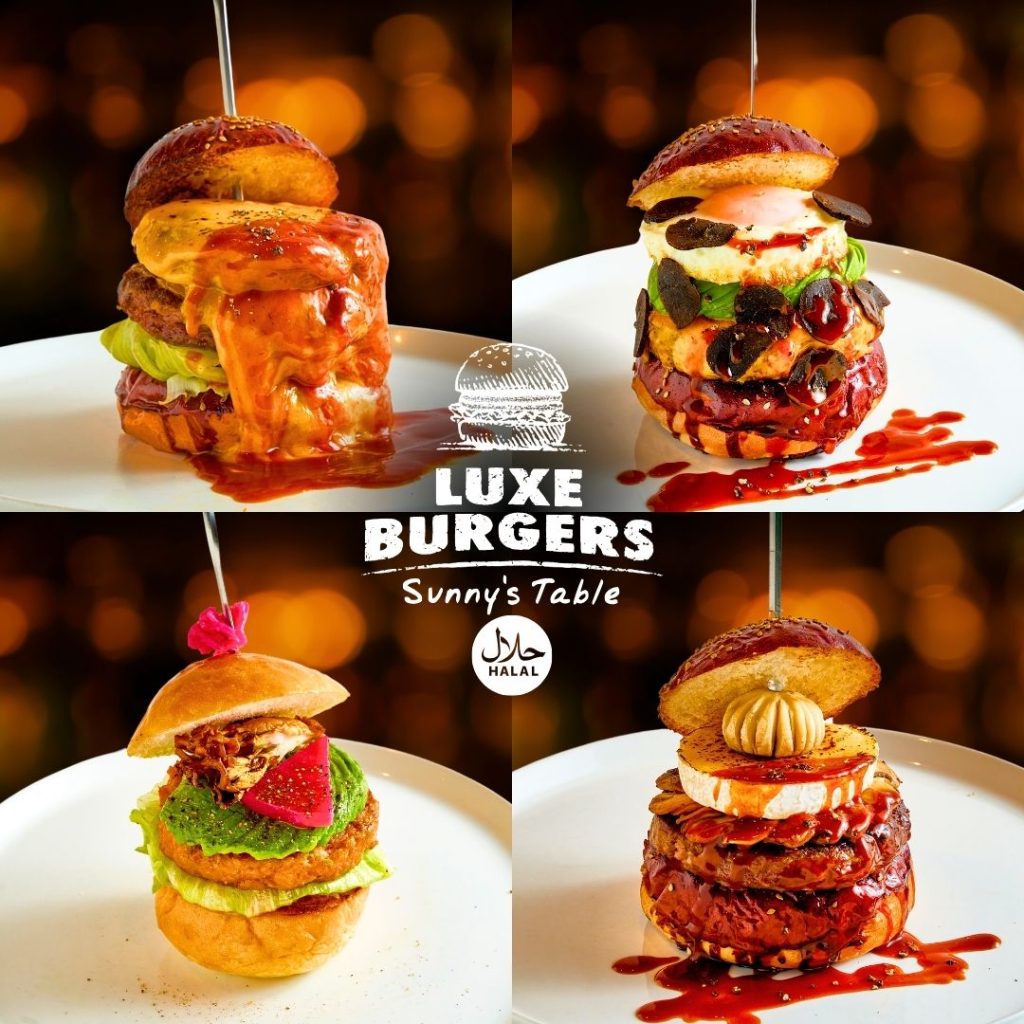 Luxe Burgers, Halal Certified, showcases four gourmet burgers. The first burger has melted cheese, mushrooms, and a fried egg on top. The second is a classic burger with cheese and lettuce. The third is a vegan burger with avocado and a pink flower garnish. The fourth burger has caramelized onions, cheese, and a truffle. Each burger is on a white plate against a wood background. The "Luxe Burgers" logo and "Sunny's Table" are in the center.